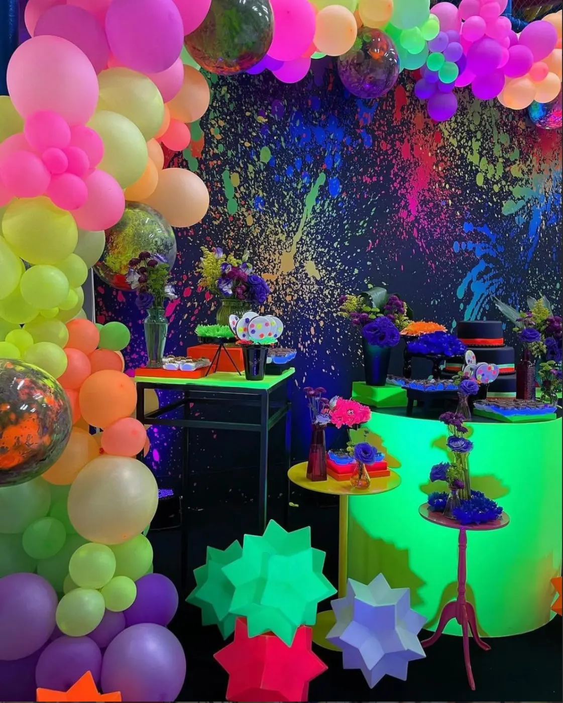 Salão de festa decorado em tema neon: mesas iluminadas, parede de respingos que brilham no escuro e arco de balões coloridos